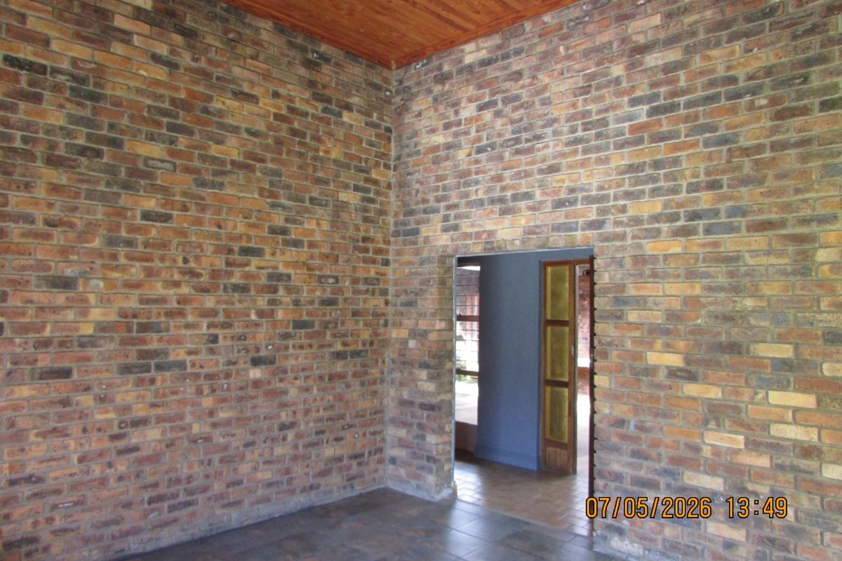 A rustic living area featuring exposed face-brick walls, tiled flooring, and a timber-paneled ceiling, leading through a doorway into an adjacent room.