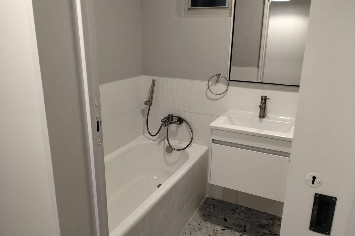 A modern, compact bathroom featuring a white bathtub with handheld shower, a wall-mounted vanity with basin, and contemporary terrazzo-style flooring.