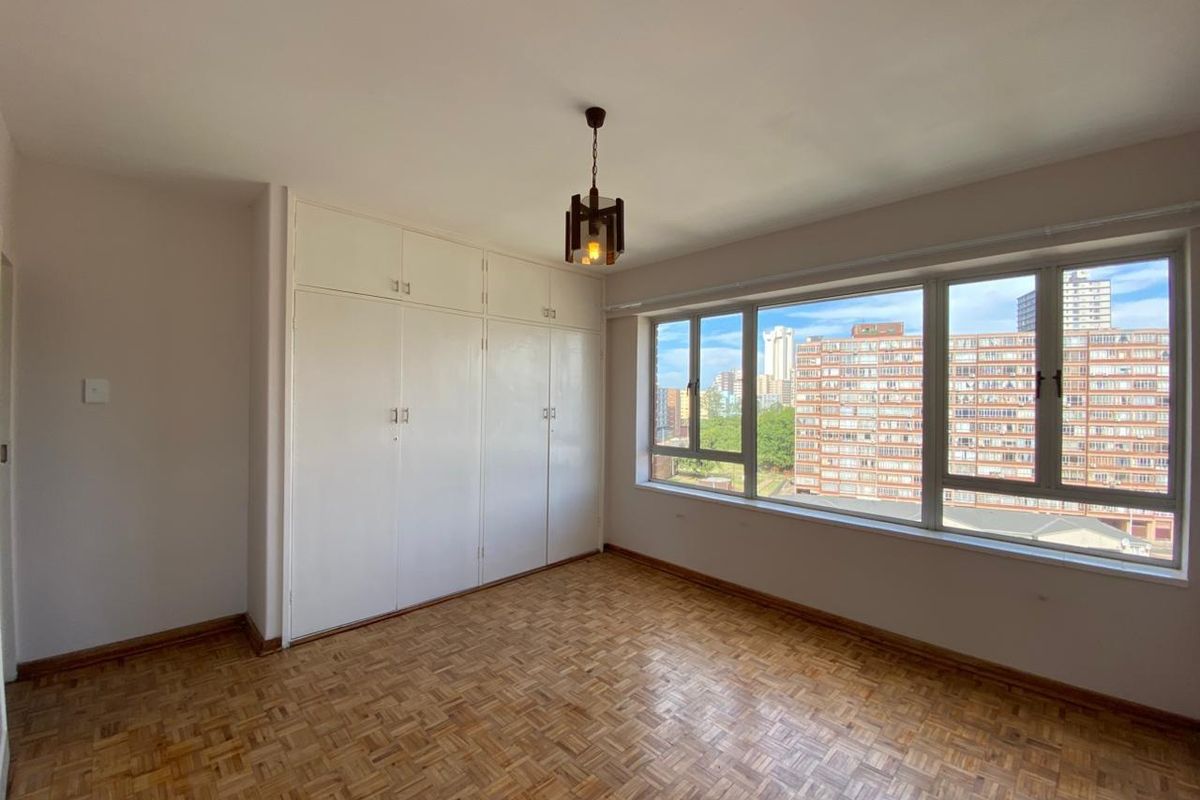 A bright, empty bedroom featuring classic parquet flooring, ample built-in cupboard storage, and large windows offering an urban city view.