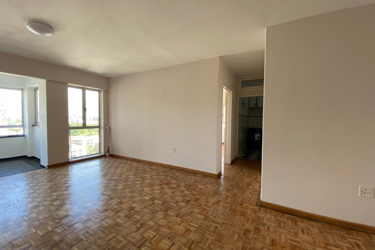 Spacious, empty living area featuring classic parquet flooring, large windows providing natural light, and neutral wall finishes.