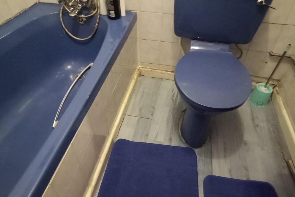 A dated bathroom featuring a blue bathtub and matching toilet, with light-colored wall tiles and wood-look flooring.