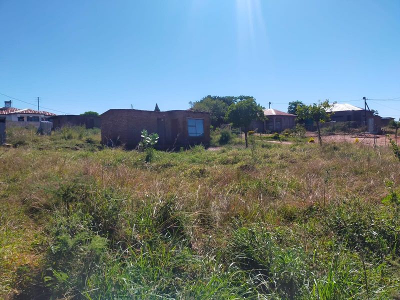 A view of a residential plot featuring an unfinished brick structure surrounded by overgrown grass and established trees in a suburban setting.