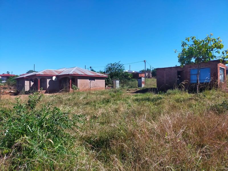 A residential plot featuring a main dwelling and a separate outbuilding structure, surrounded by undeveloped land with natural vegetation.