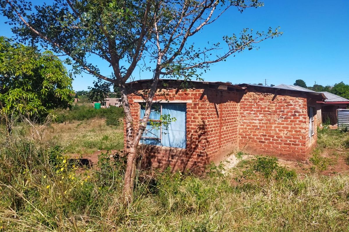 A view of a small, basic brick structure situated on a vacant residential plot, showing signs of age and requiring maintenance.