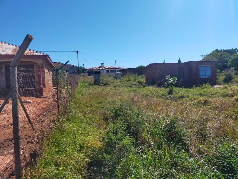 A view of a residential plot featuring overgrown grass, existing boundary fencing, and basic outbuildings on the property.