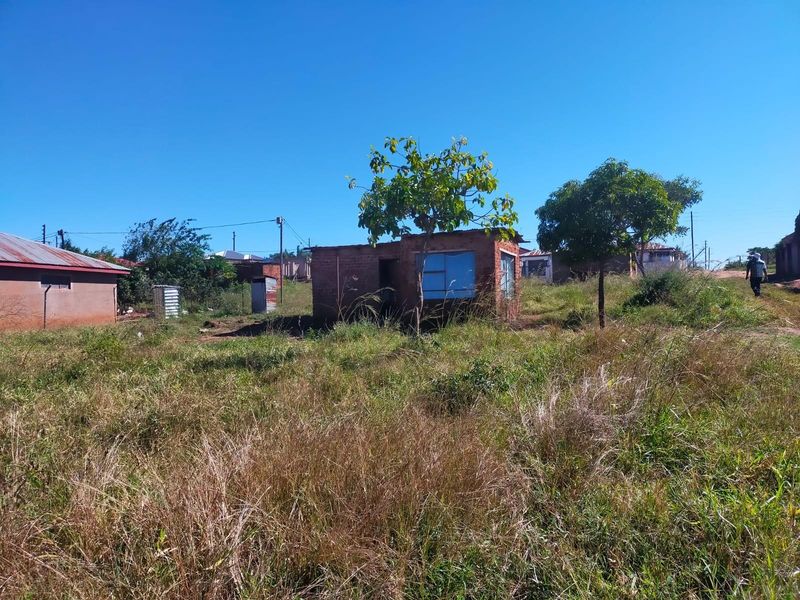 A view of a residential plot featuring a small, basic brick structure surrounded by overgrown grass and mature trees.