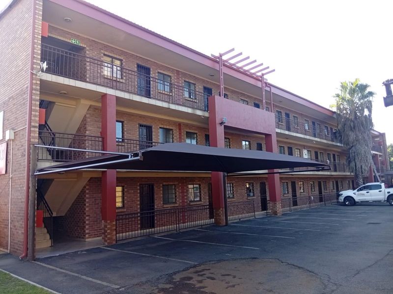 Exterior view of a multi-story apartment building featuring face brick construction, metal balustrades, and a covered parking area with shade cloth.