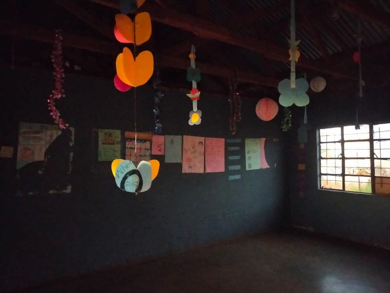 An interior space featuring exposed timber roof beams, a corrugated metal ceiling, and a steel-framed window, currently used as a classroom or utility area.