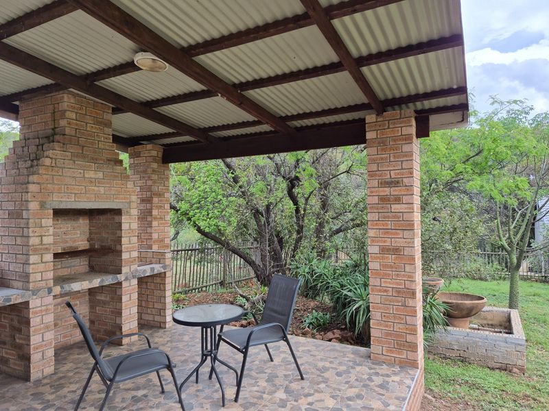 A covered outdoor patio featuring a built-in brick braai or fireplace, exposed timber roof structure, and tiled flooring overlooking a garden.