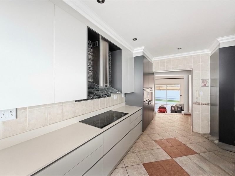 Modern galley-style kitchen featuring sleek cabinetry, integrated cooktop, stainless steel extractor fan, and tiled flooring, leading through to an adjacent living area.