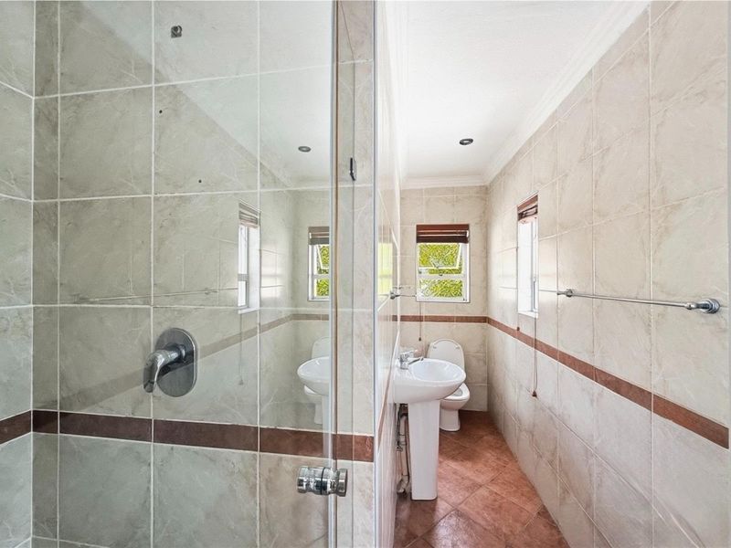 A functional bathroom featuring a glass-enclosed shower, pedestal basin, and toilet with neutral wall tiling and contrasting floor tiles.