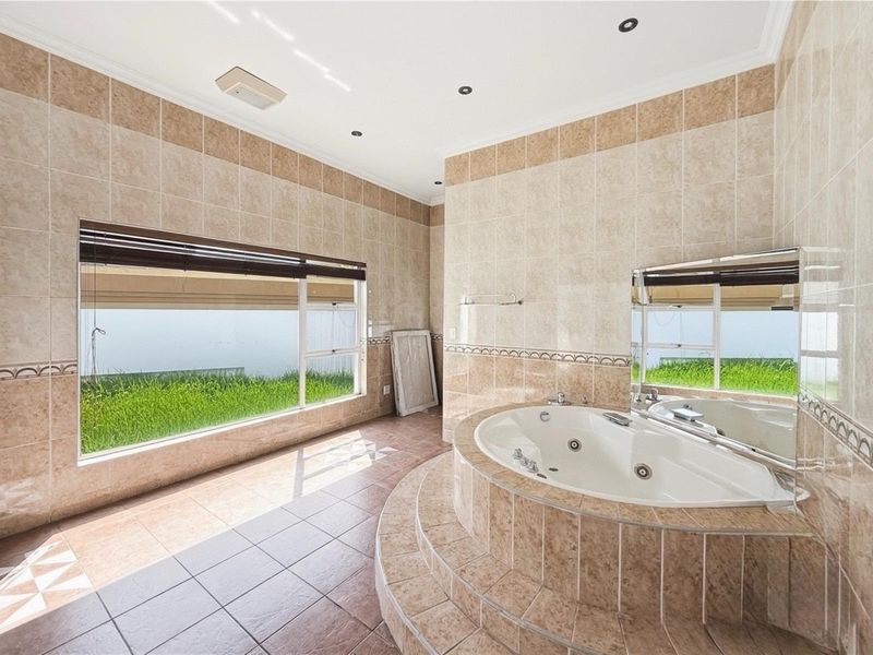 Spacious bathroom featuring a raised corner spa bath, floor-to-ceiling neutral tiling, and large windows overlooking a garden area.