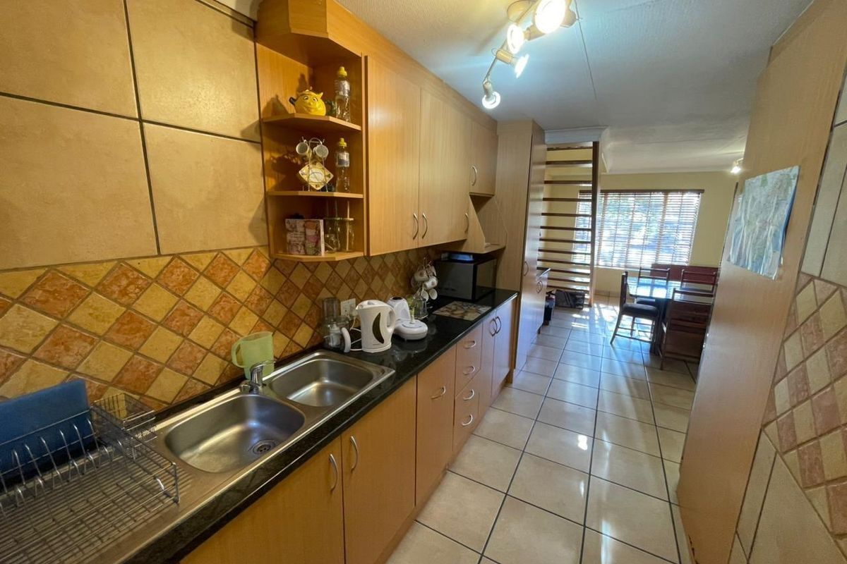 A functional galley-style kitchen featuring wood-finish cabinetry, dark countertops, double stainless steel sink, and tiled flooring, leading into an open-plan dining area.