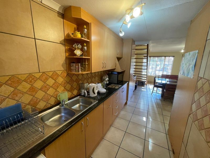 A functional galley-style kitchen featuring wood-finish cabinetry, dark countertops, double stainless steel sink, and tiled flooring, leading into an open-plan dining area.