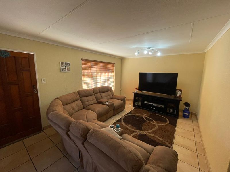 A living room featuring tiled flooring, a wooden entrance door, and window blinds, providing a functional and comfortable space for relaxation.