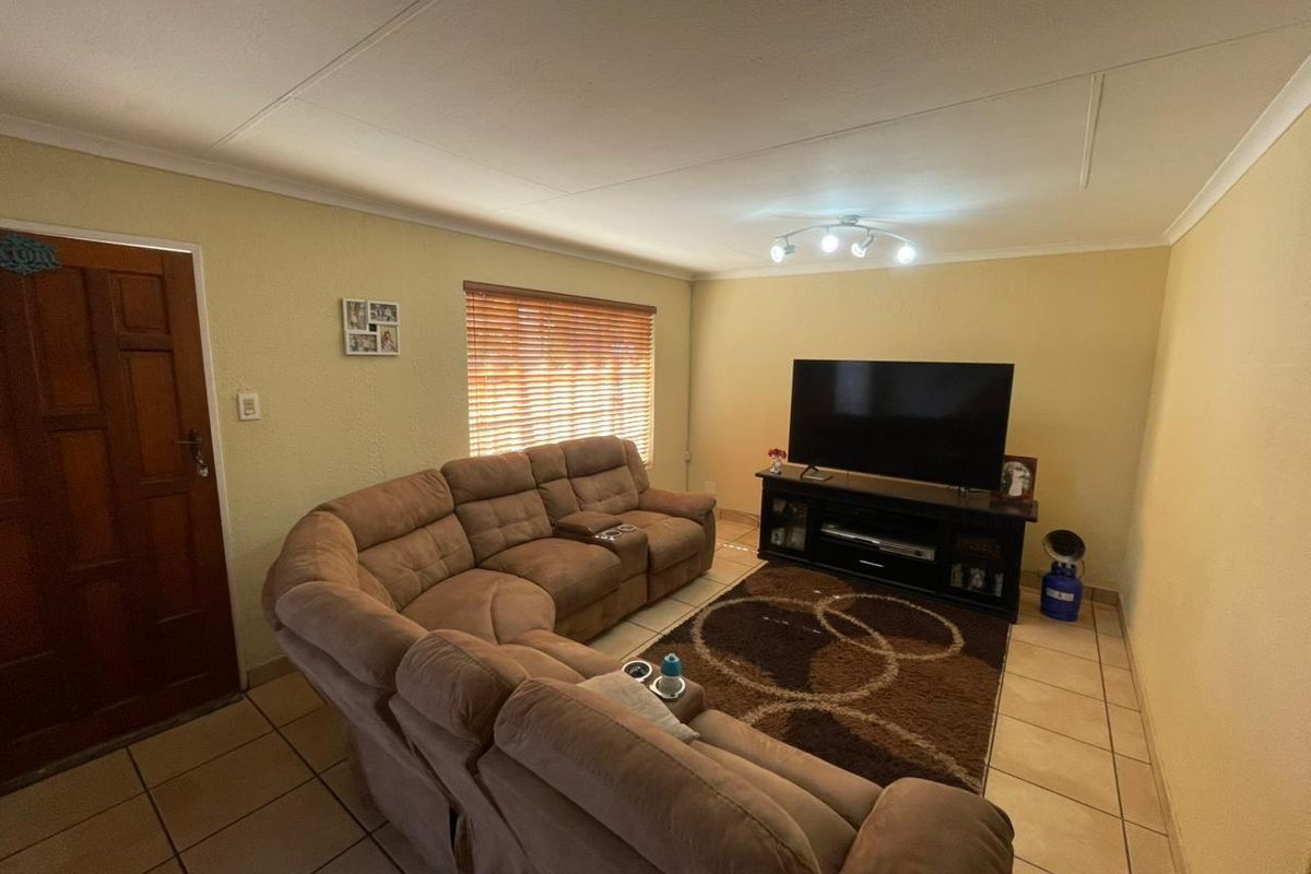 A living room featuring tiled flooring, a wooden entrance door, and window blinds, providing a functional and comfortable space for relaxation.