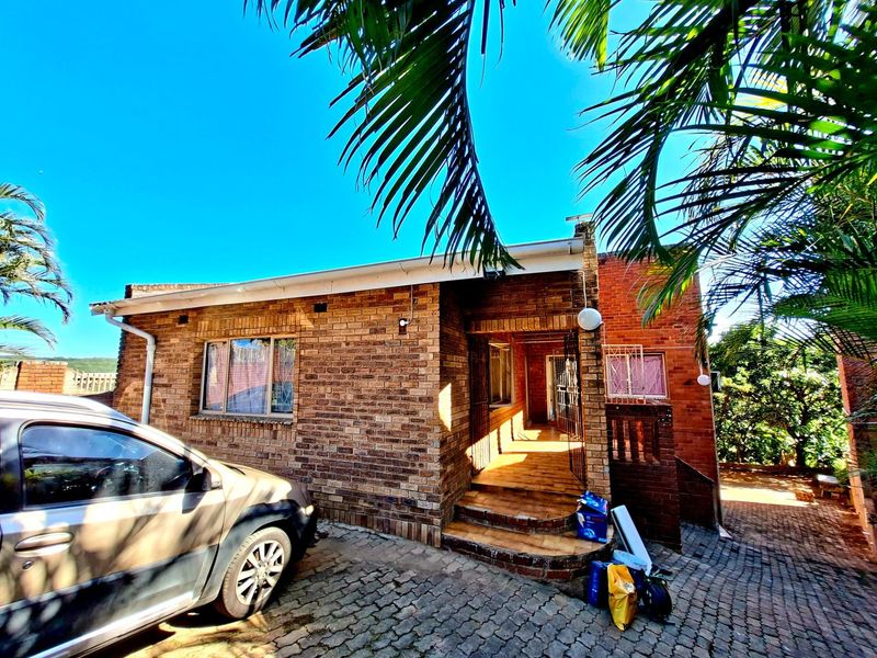 A front exterior view of a face-brick house featuring a paved driveway, covered entrance porch, and established palm trees.