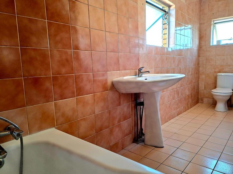 A dated bathroom featuring full-height terracotta-toned wall tiles, a white pedestal basin, a standard toilet, and a bathtub with a handheld shower attachment.