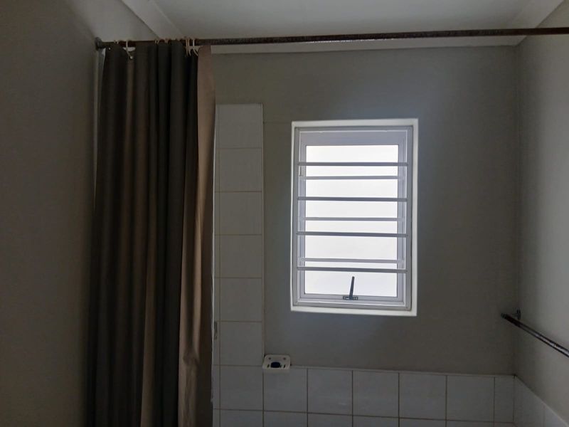 A functional bathroom space featuring a tiled splashback, window with security bars, and basic shower fixtures.