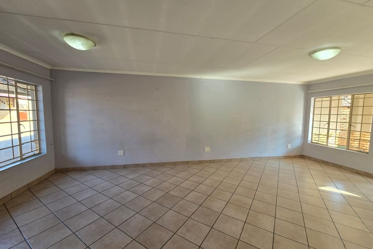 A spacious, empty living room featuring neutral tiled flooring, large windows with security bars, and dual ceiling-mounted light fixtures.