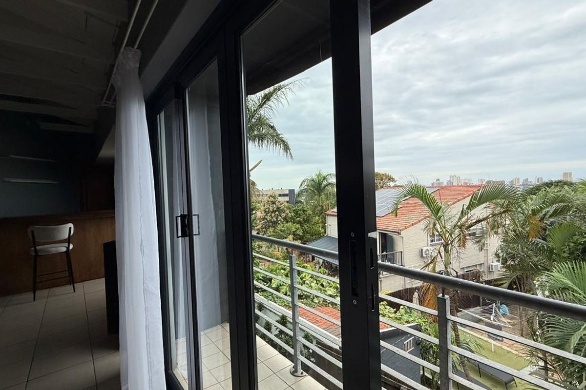 A view from an interior living space through large sliding glass doors onto a balcony overlooking a suburban garden and neighbourhood.