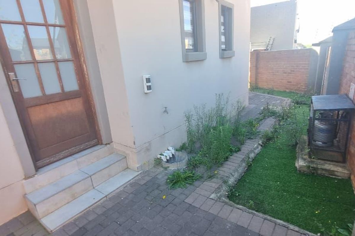 A rear exterior view of the property featuring a paved walkway, small garden patch, and access door with concrete steps.