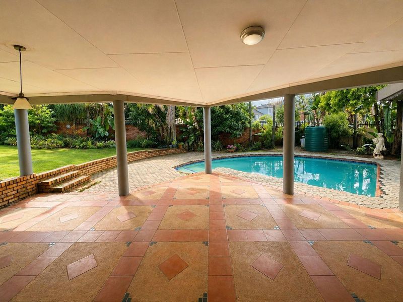 Spacious covered patio overlooking a swimming pool and landscaped garden, featuring tiled flooring and structural support columns.