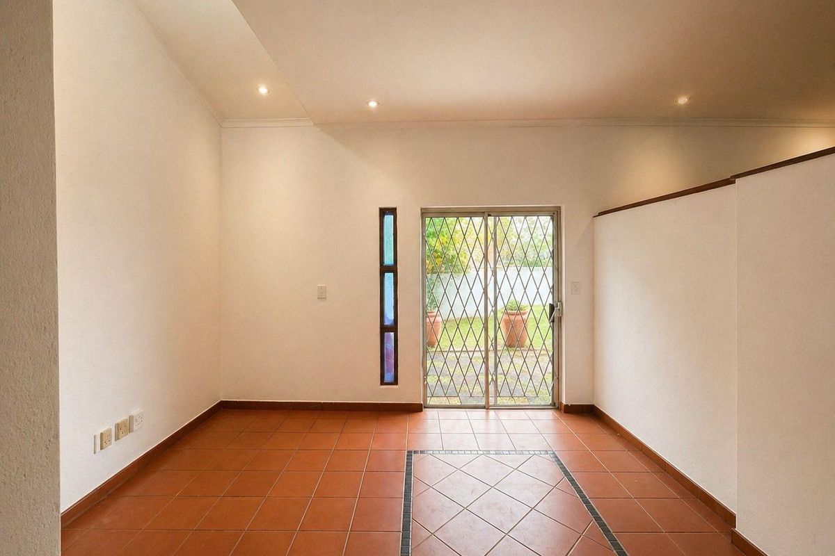 A bright, empty living area featuring terracotta floor tiles, recessed ceiling lighting, and a sliding glass door with security gate leading to the garden.