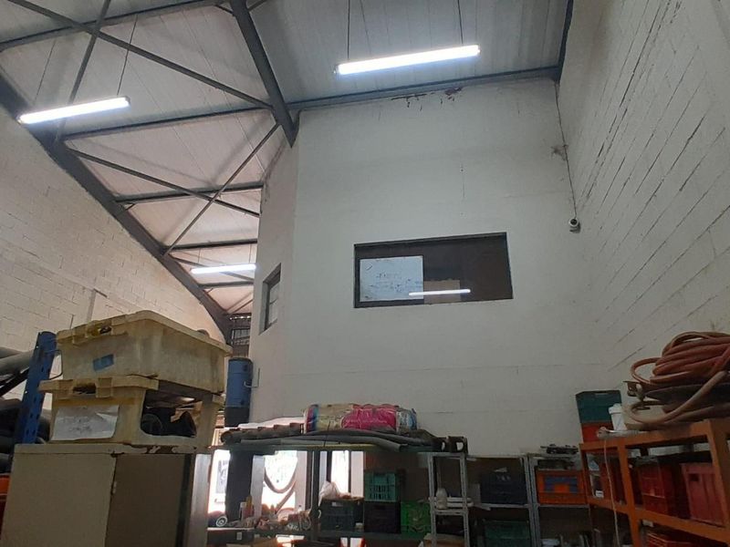 Industrial warehouse space featuring high ceilings with exposed steel trusses, fluorescent lighting, and a mezzanine-level office window overlooking the main floor area.