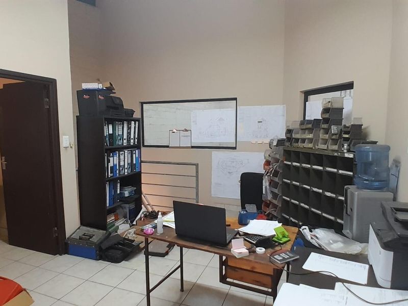 Functional industrial office space featuring tiled flooring, internal window partition, and direct access to a small washroom facility.