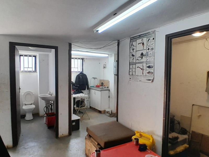 Industrial utility area featuring a small bathroom, kitchenette prep space, and multiple access points with basic fluorescent lighting and concrete flooring.