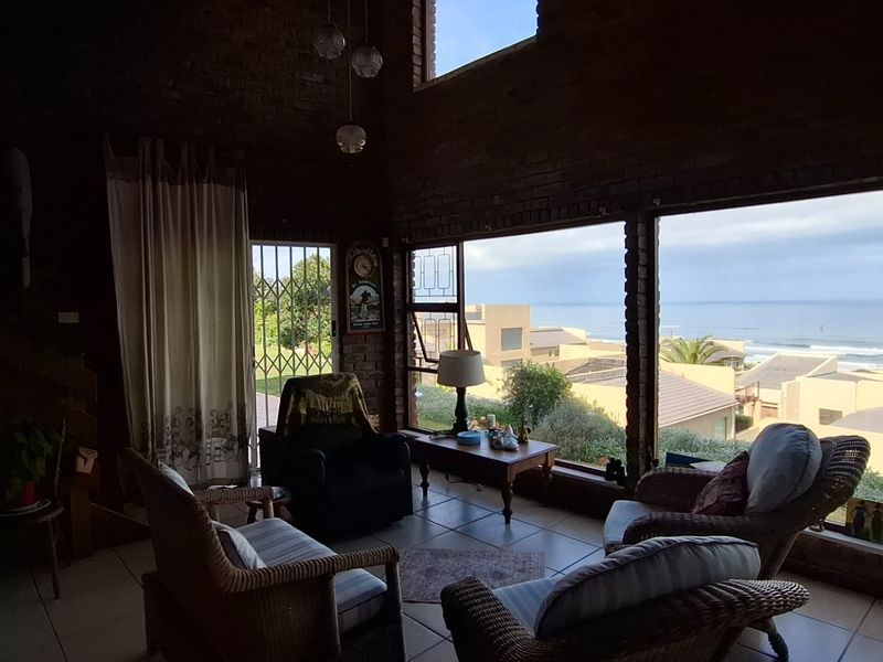 Spacious living area featuring double-volume exposed brick walls, large windows with ocean views, and tiled flooring, providing a bright and airy coastal atmosphere.