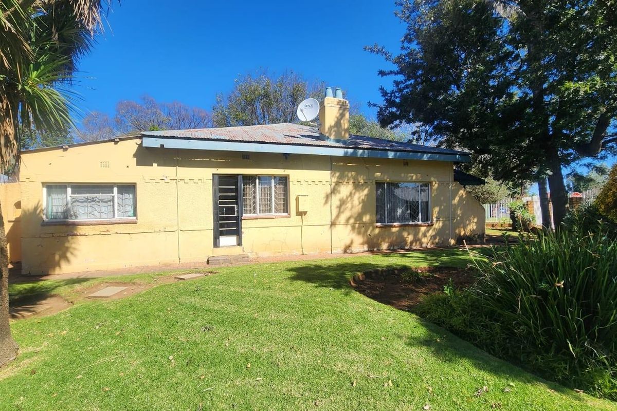 A single-story residential house featuring a yellow stucco facade, corrugated metal roof, and a surrounding garden area.