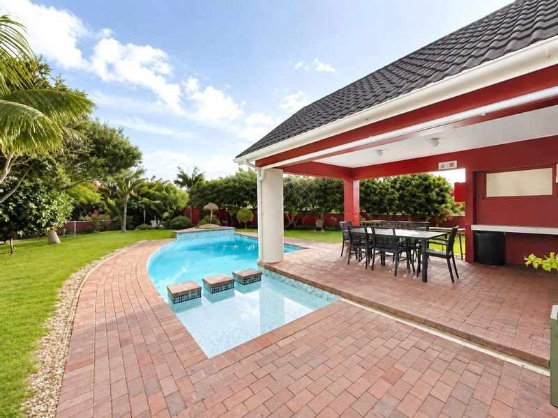 Outdoor entertainment area featuring a covered patio with built-in counter, brick paving, and a swimming pool set within a landscaped garden.