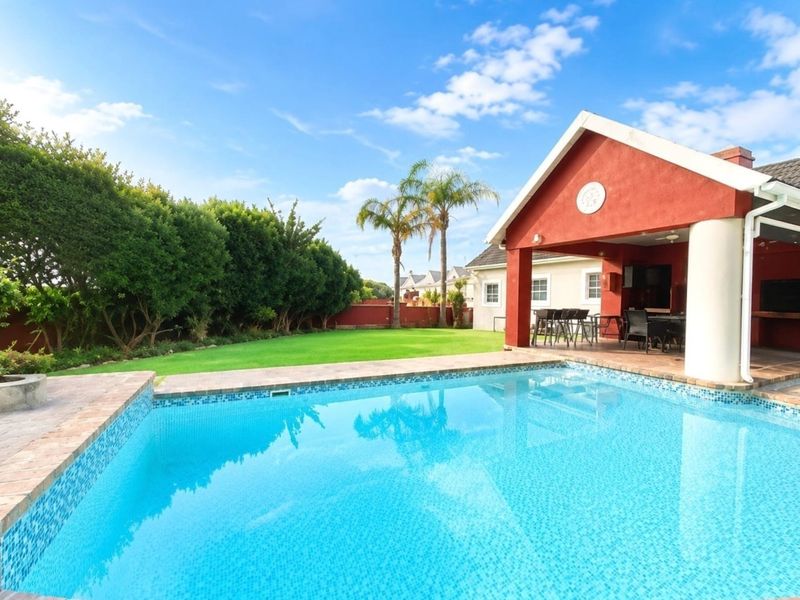 A well-maintained outdoor area featuring a sparkling swimming pool, a spacious covered patio with brick pillars, and a lush, manicured garden.