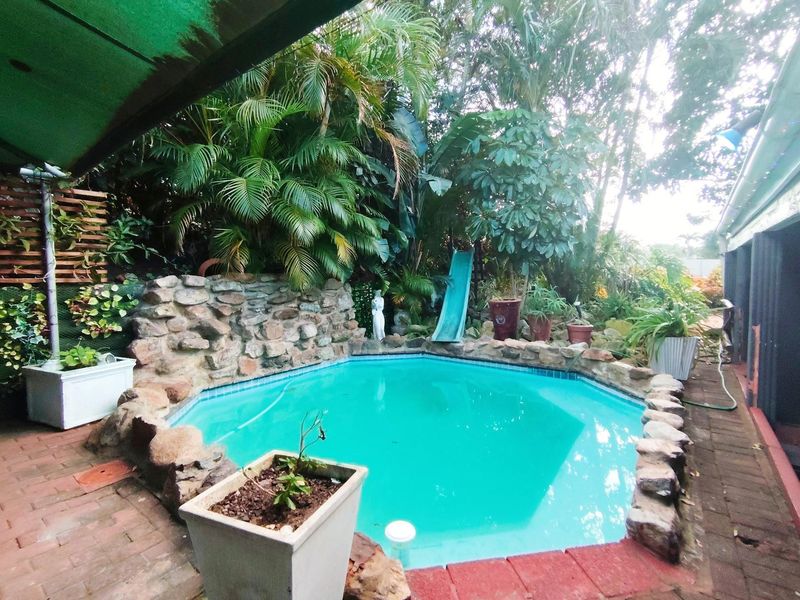 A backyard swimming pool area featuring a stone rockery surround, a built-in slide, and brick paving, set within a lush, established garden.