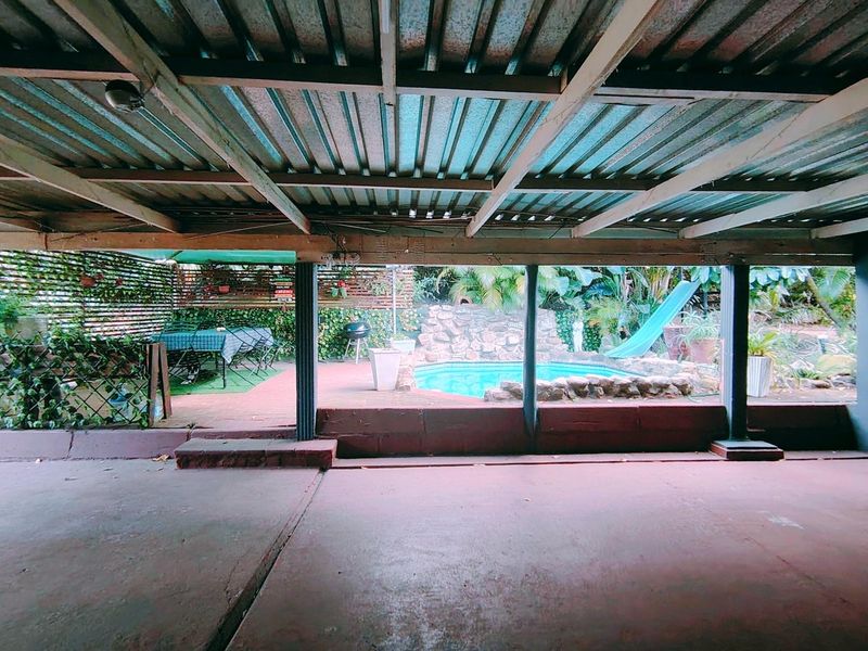 Covered outdoor patio area featuring a concrete floor and timber structure, overlooking a private swimming pool with stone surround and slide.