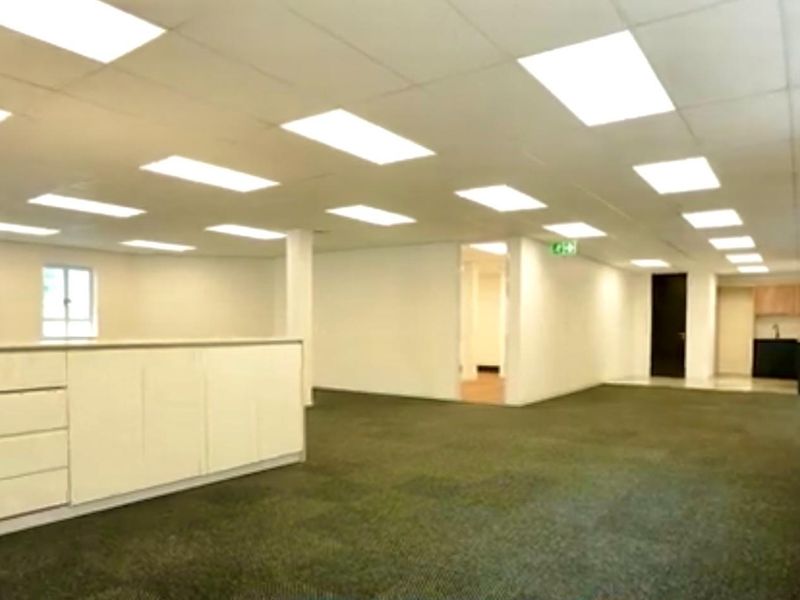 Spacious open-plan commercial office area featuring recessed ceiling lighting, neutral finishes, and a small kitchenette area in the background.