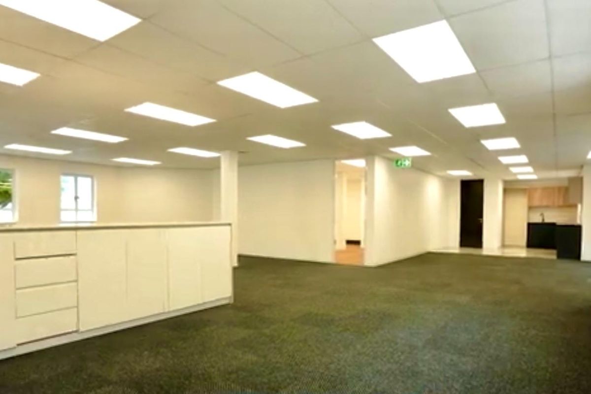 Spacious open-plan commercial office area featuring recessed ceiling lighting, neutral finishes, and a small kitchenette area in the background.