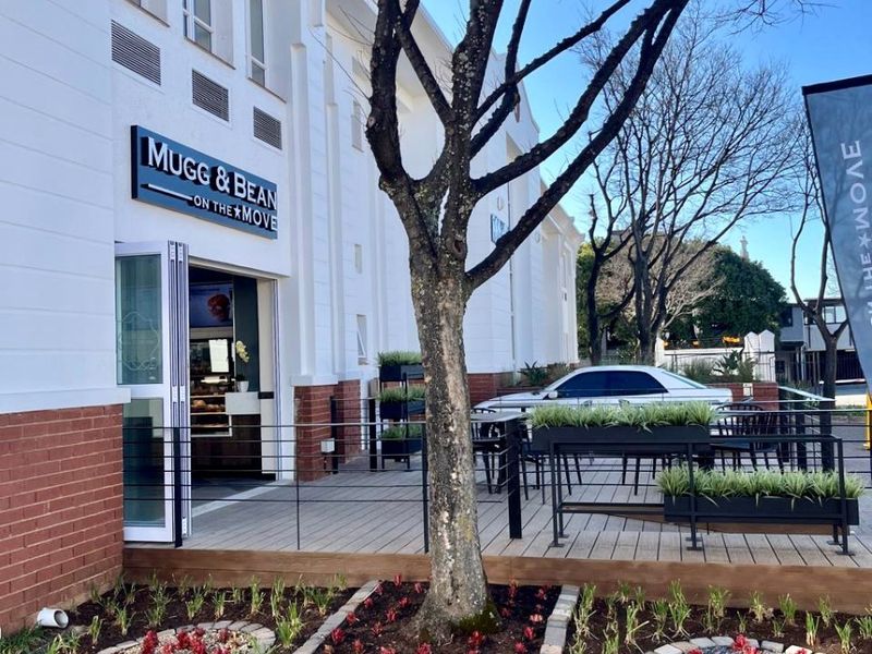 Exterior view of a commercial storefront featuring a wooden deck patio, landscaped garden beds, and modern building facade with signage.