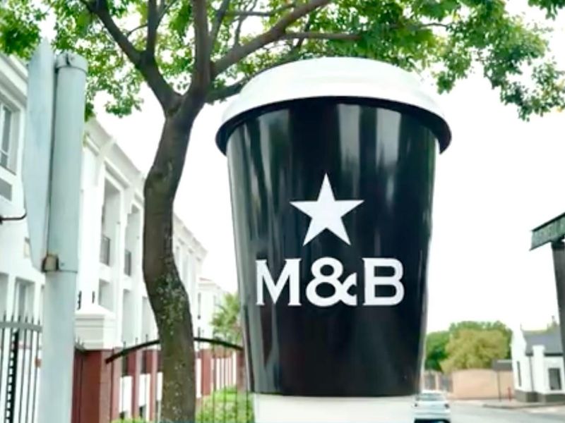 Exterior view of a commercial property featuring a prominent M&B branded coffee cup signage and surrounding streetscape.