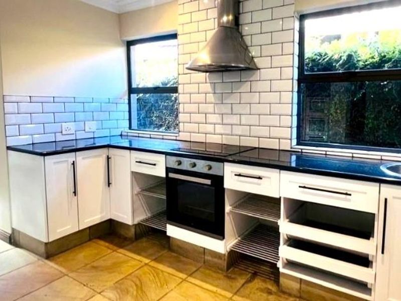 A modern kitchen featuring white cabinetry, subway tile backsplash, stainless steel extractor fan, built-in oven, and dark countertops with a stainless steel sink.