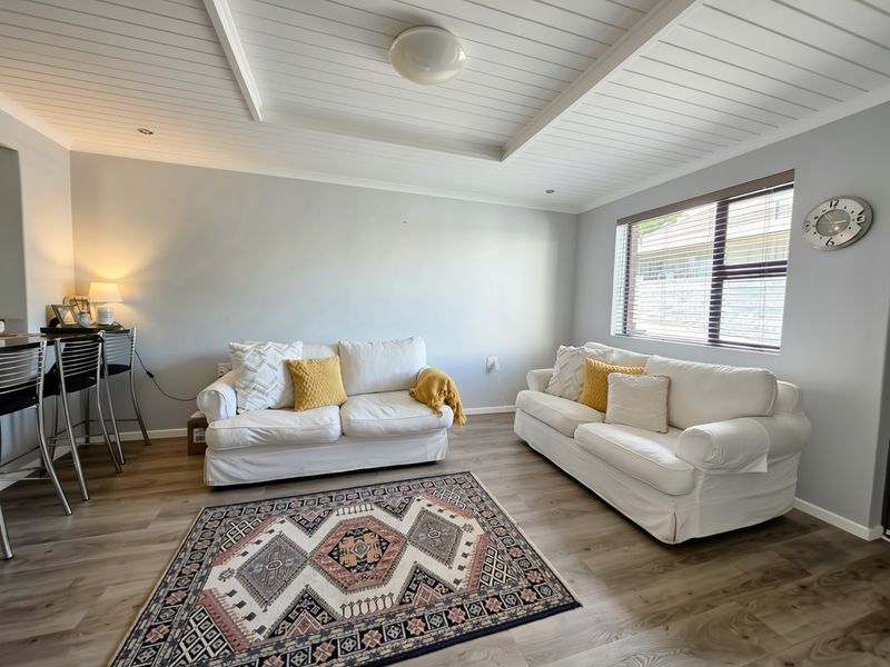 A bright and well-maintained living room featuring modern laminate flooring, a classic tongue and groove ceiling, and large windows with fitted venetian blinds.