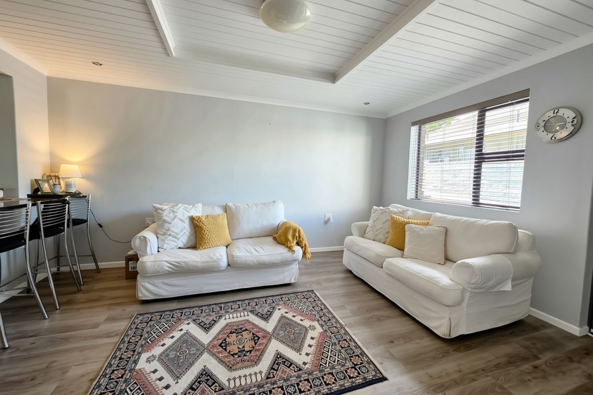 A bright and well-maintained living room featuring modern laminate flooring, a classic tongue and groove ceiling, and large windows with fitted venetian blinds.