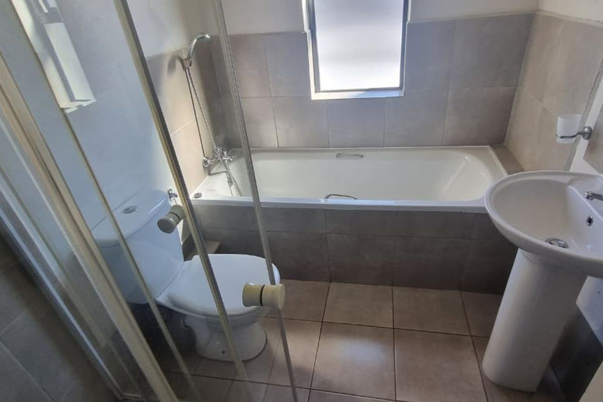 A functional bathroom featuring a tiled bathtub, separate glass-enclosed shower, pedestal basin, and toilet with neutral wall and floor tiling.