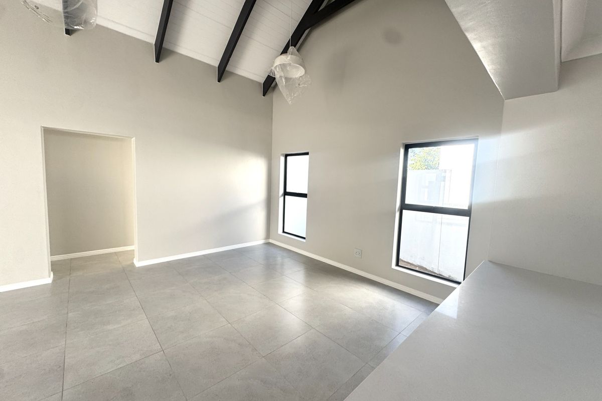 A bright, modern living area featuring high vaulted ceilings with exposed beams, large windows, and neutral porcelain tile flooring in a newly finished space.