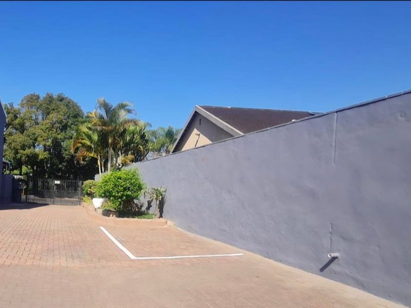 A paved driveway area featuring a long boundary wall, gated access, and established perimeter landscaping within a townhouse complex.