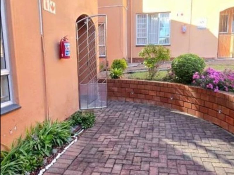 A paved courtyard area featuring a low brick retaining wall, landscaped garden beds, and a security gate entrance to the townhouse.
