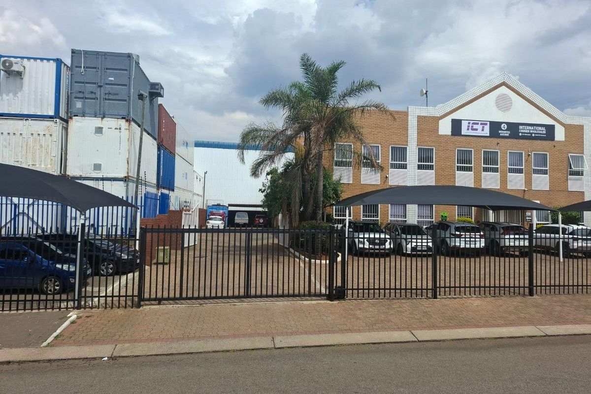 Front view of an industrial property featuring a brick facade, paved driveway, secure metal fencing, and shade cloth parking structures.