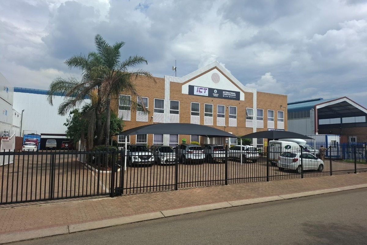 Front view of an industrial warehouse facility featuring a brick facade, paved parking area with shade cloth carports, and secure perimeter fencing.
